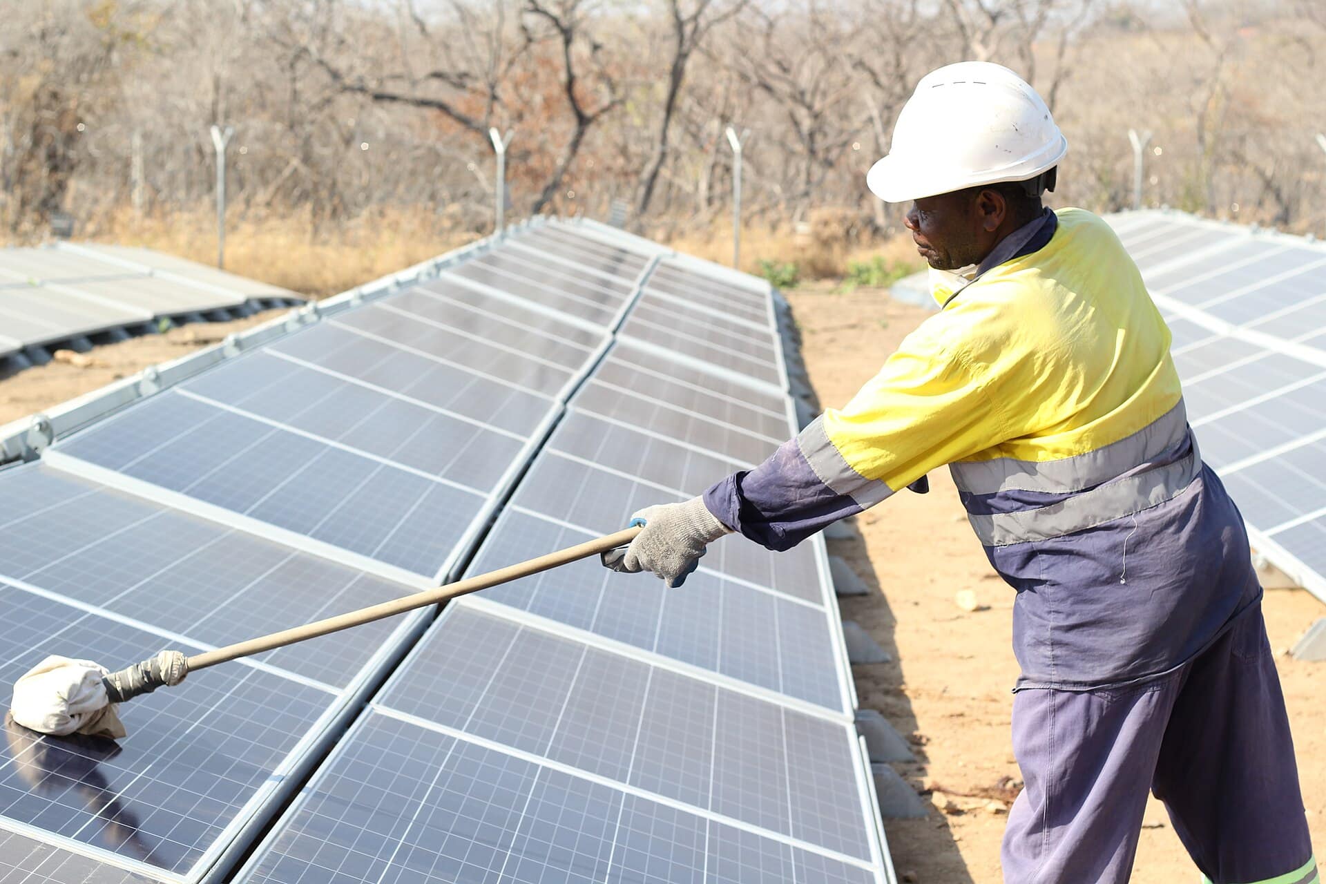 Worker in a hard hat cleaning a rooftop solar panel with a long-handled brush