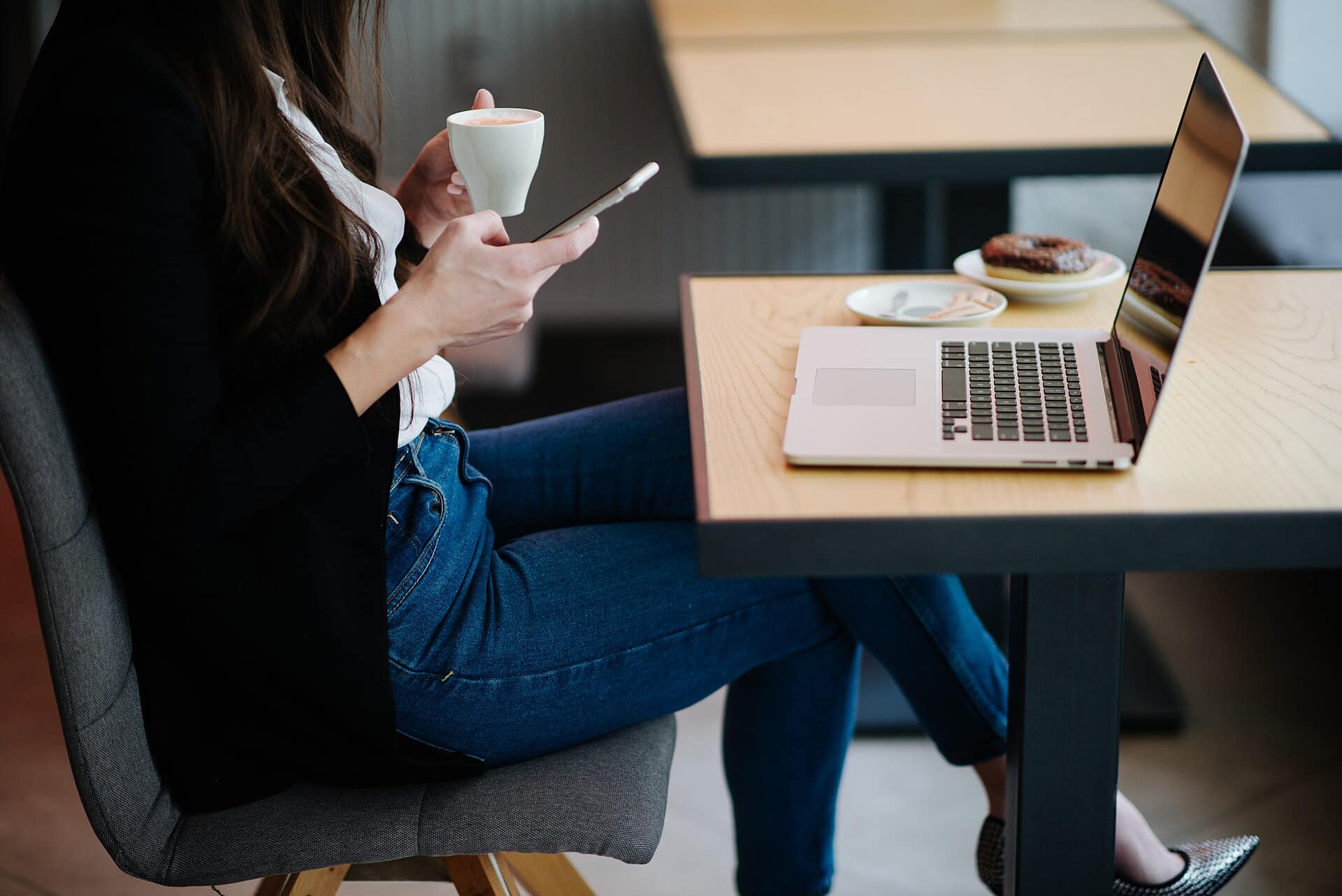 Person with a phone seated at a cafe table beside an open laptop and a cup of coffee