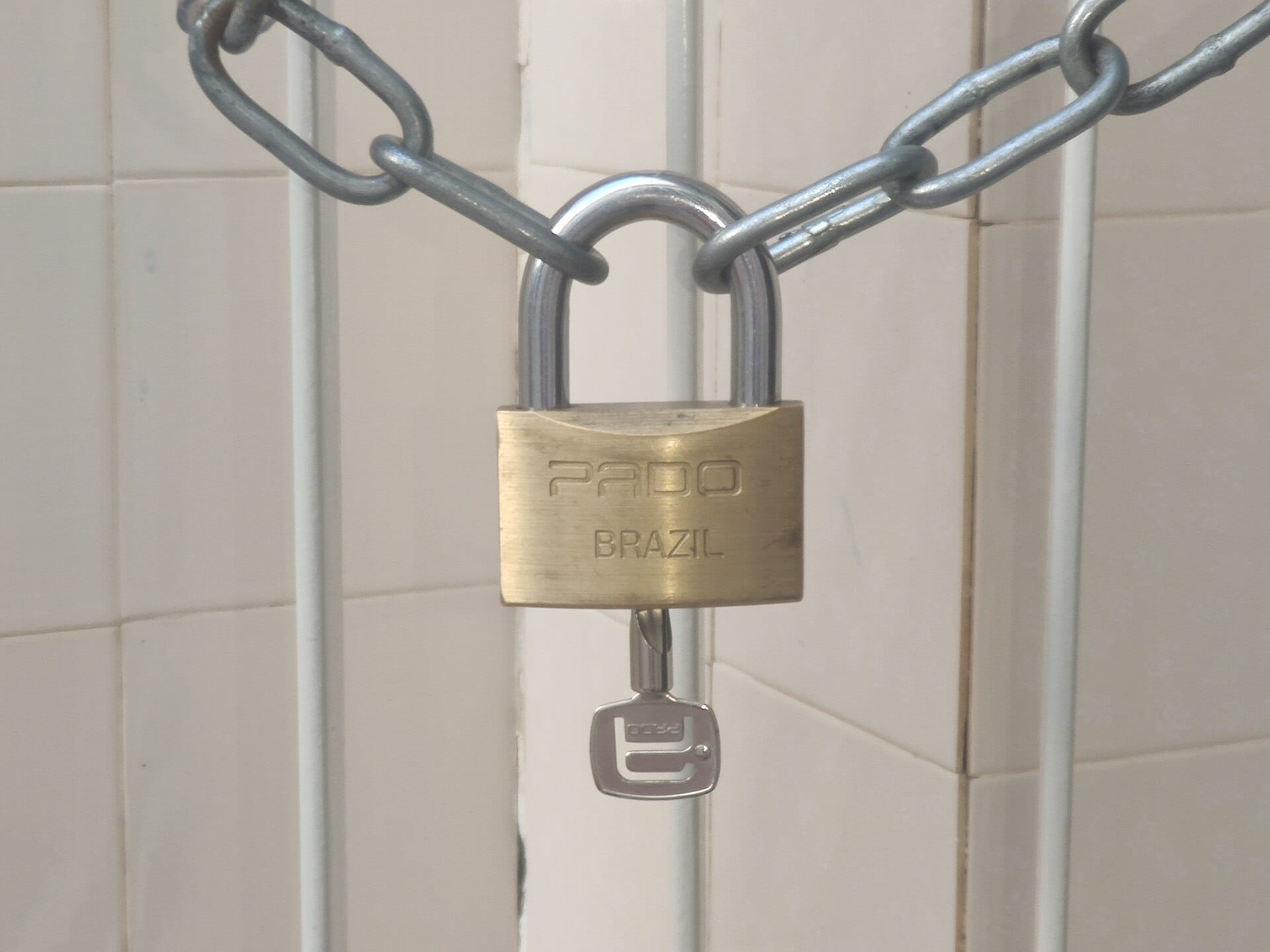 Brass padlock hanging from a steel chain against a tiled wall